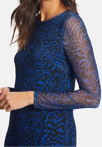 Žena v dlouhém rukávu, průhledném topu modro-černého vzoru se vířivými abstraktními motivy, který je zkrácený pod rameny a nad pasem.