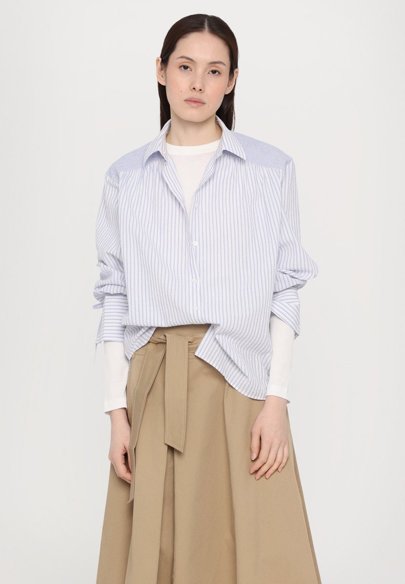 Chemise boutonnée rayée bleue et blanche à manches retroussées, associée à une jupe beige taille haute avec une ceinture nouée.