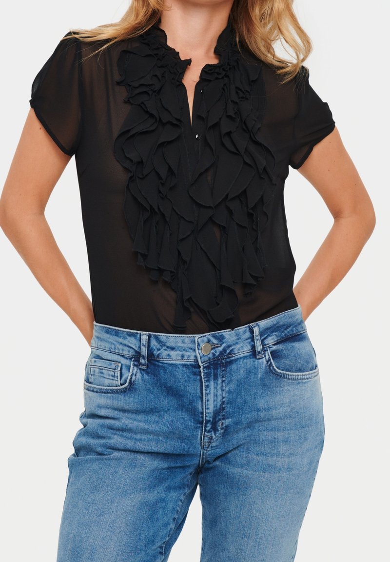 Blusa nera trasparente con dettaglio arricciato sul davanti, maniche corte e scollo rotondo, abbinata a jeans di jeans blu con vestibilità classica.