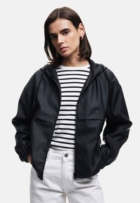 Zwarte hooded jacket met een ritssluiting aan de voorkant, met elastische manchetten, een gladde textuur, gedragen over een wit en zwart gestreept shirt met een ronde hals.