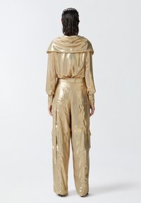 Zlatni metalik top s drapiranim ovratnikom, uparen s odgovarajućim cargo hlačama koje imaju džepove i glatku, sjajnu teksturu.