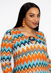 Bunt gemusterte Bluse mit Zickzack-Muster in Orange, Türkis und Schwarz. Verfügt über einen Ausschnitt mit Schlüsselloch und strukturierter Strickstoff. Kombiniert mit orangen Ohrringen.