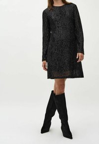 Robe noire à manches longues avec un motif texturé en sequins et une coupe ample. Associée à des bottes en daim noires montant jusqu'aux genoux avec des bouts pointus.