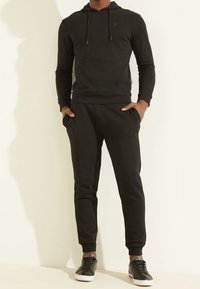 Persoon in een zwarte hoodie en bijpassende joggingbroek met handen in de zakken, staande tegen een effen lichte achtergrond.