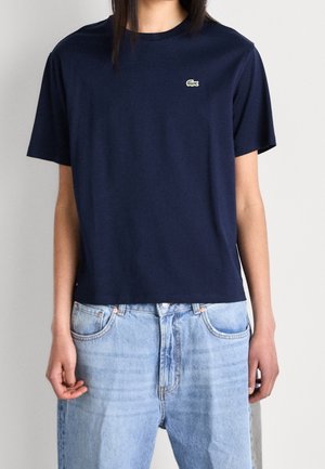 Personne portant un t-shirt Lacoste bleu marine à manches courtes et un jean taille haute bleu clair, debout devant un fond clair uni.