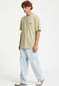 Lysegrøn oversized t-shirt med korte ærmer og en lille sort logo, kombineret med løstsiddende lyseblå jeans og brune sportssko.