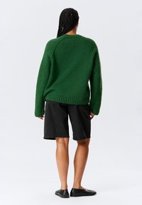 Persoon met lang gevlochten haar, gekleed in een gestructureerde groene trui, zwarte knielange shorts en zwarte instappers, van achteraf gezien op een witte achtergrond.