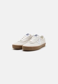 Beige suede sneakers with white accents, a rounded toe, flat tan rubber sole, and beige laces. Features a logo tag on the tongue.