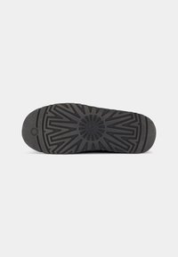 Black rubber shoe sole with a geometric pattern, featuring pronounced ridges and a circular logo at the center. Durable texture for traction.