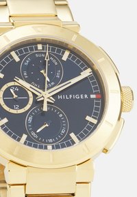 Gullfarget rustfritt stålklokke med svart urskive, flere sub-skiver og datovisning; har en teksturert krone og et glatt metallarmbånd.