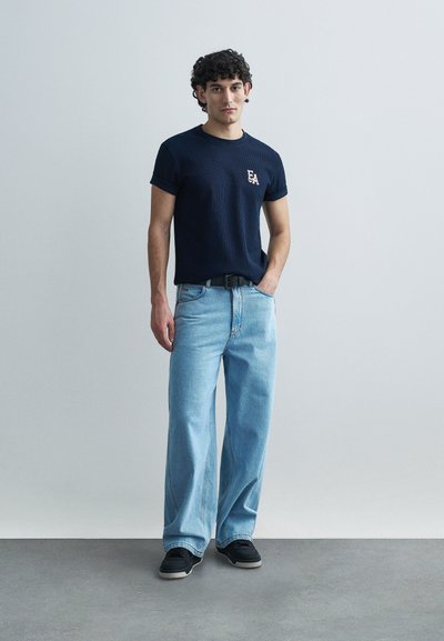 Μπλε ναυτικό T-shirt με κοντά μανίκια και ριμπ υφή, διακοσμημένο με λογότυπο, συνδυασμένο με φαρδιά τζιν παντελόνια ανοιχτού μπλε χρώματος και μαύρα αθλητικά παπούτσια. Απλό, καθημερινό ντύσιμο.