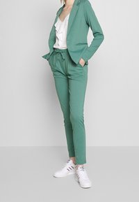 Grön, figursydd kavaj med matchande byxor, tillverkad av ett slätt tyg. Vit rufflad topp under. Vita sneakers med färgade ränder.