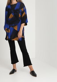 Blå og orange mønstret kimono-stil top med brede ærmer, parret med sorte flarebukser og sorte slip-on sko. Glat stoftekstur.