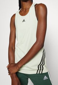 Débardeur de sport vert clair avec un logo Adidas noir, présentant une texture lisse, un dos nageur et des bandes latérales noires, associé à un pantalon vert.