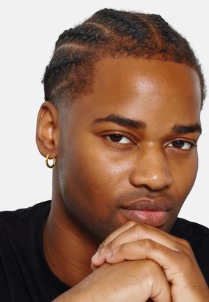 Boucle d'oreille en or, texturée, sur l'oreille d'une personne. Gros plan du visage avec une coiffure tressée soignée, une peau lisse et des mains jointes devant.