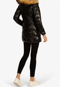 Manteau noir matelassé avec un motif en chevrons, doté d'une capuche en fourrure. Porté sur des leggings noirs ajustés et des baskets blanches.