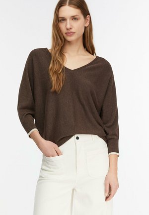 Kobieta z długimi włosami, ubrana w luźny brązowy sweter z dekoltem w serek i białe spodnie z wysokim stanem, z jedną ręką w kieszeni.