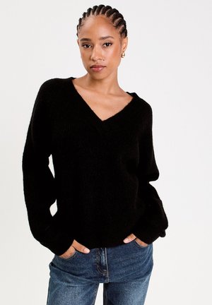 Schwarzer Strickpullover mit V-Ausschnitt und langen Ärmeln, getragen über blauen Denim-Jeans. Der Stoff wirkt weich mit einer leicht strukturierten Oberfläche.