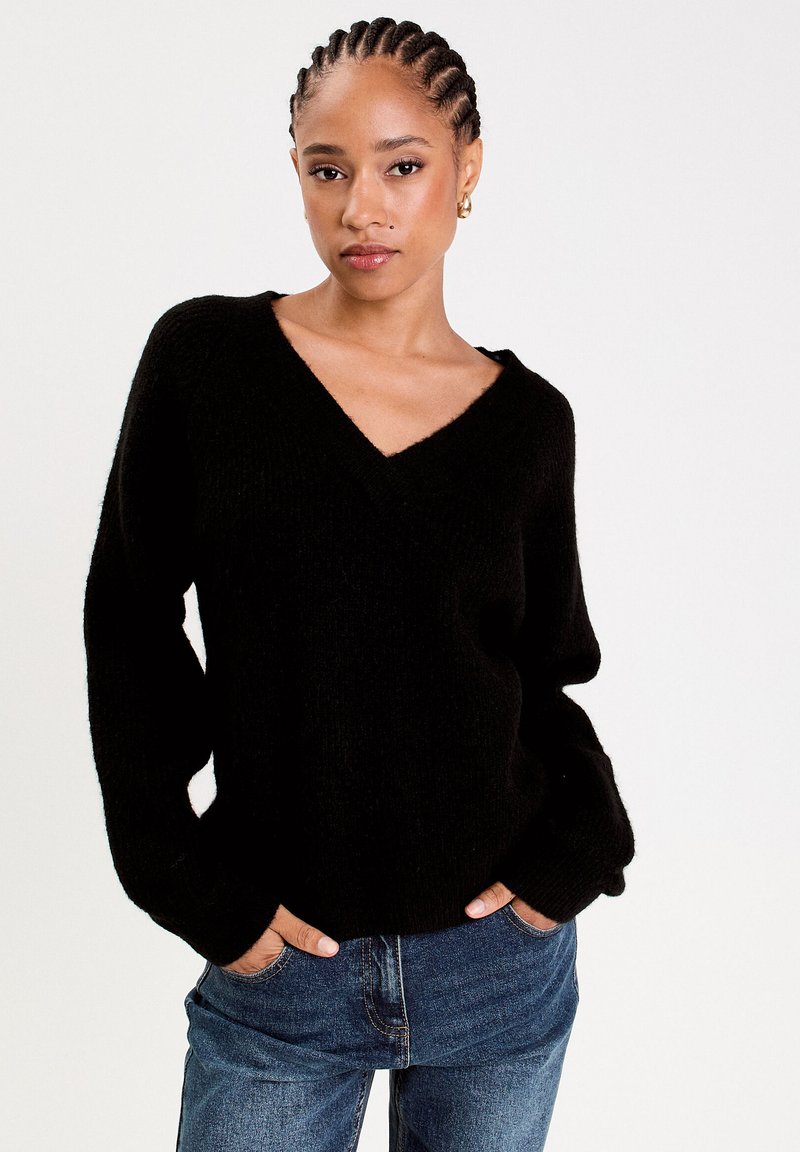 Schwarzer Strickpullover mit V-Ausschnitt und langen Ärmeln, getragen über blauen Denim-Jeans. Der Stoff wirkt weich mit einer leicht strukturierten Oberfläche.