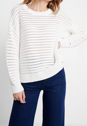 Mujer con un suéter blanco de punto abierto y vaqueros azul oscuro de talle alto, con una mano en la cadera y la otra relajada.