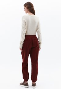 Béžový top s dlouhým rukávem, směs bavlny. Červené kordové kalhoty, elastický pas, zúžené manžety. Model má na sobě hnědé tenisky. Bílý pozadí.