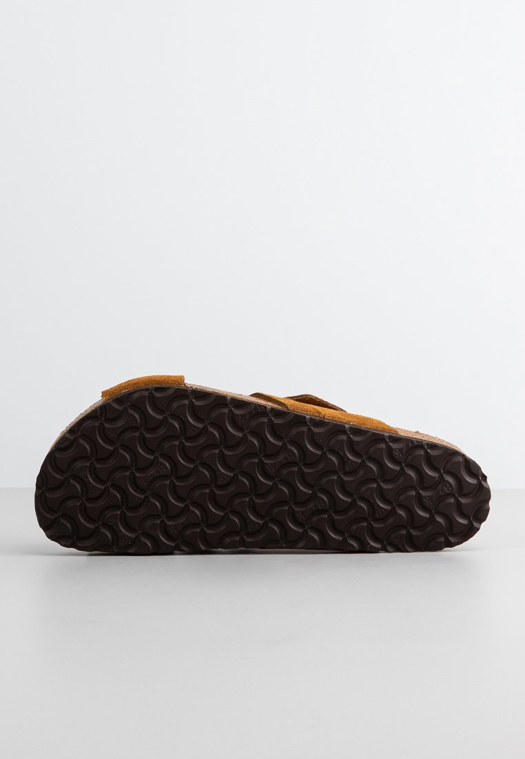 Sandal en daim marron avec une semelle en caoutchouc noir texturé, dotée d'un design de courroie simple pour un ajustement secure et un confort.