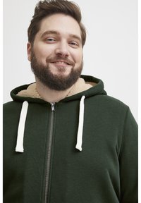 Sudadera con cremallera de color verde oscuro con forro de sherpa beige, cordones blancos y cremallera frontal. Textura de tela suave.