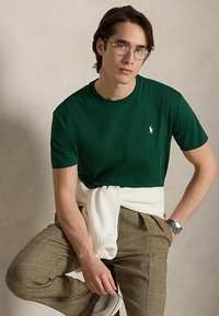 Jeune homme portant un t-shirt vert, un pantalon à carreaux, un pull blanc noué à la taille, des lunettes et une montre, assis avec un genou relevé sur un fond uni.