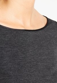 Černý, strukturovaný oděv s širokým výstřihem a kontrastním lemováním. Měkký materiál s jemným melírovaným vzorem. Detailní pohled na oblast límce.