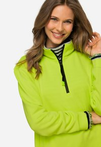 Sudadera de polar verde lima brillante con cremallera de cuarto de vuelta en el frente, cremallera negra y cuello a rayas. Los puños tienen un borde rizado. Textura suave.
