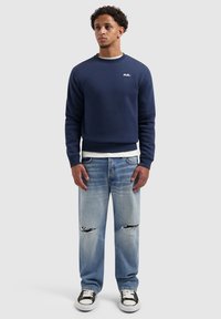 Man in een marineblauwe sweatshirt, witte t-shirt, lichte blauwe ripped jeans en zwart-witte sneakers, staand tegen een effen achtergrond.
