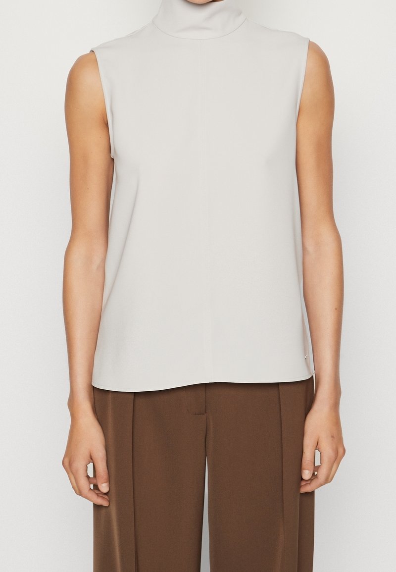 Haut sans manches à col roulé en tissu beige clair, présentant une coupe droite et des coutures nettes, associé à un pantalon à jambes larges marron.