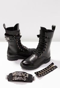 Bottes montantes en cuir noir avec bouts ronds, œillets métalliques et sangles texturées. Comprend une sangle décorative avec l'inscription « VERSACE JEANS » en lettres argentées.