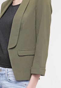 Olivově zelený blazer s texturovaným povrchem, klopami a srolovanými rukávy, v kombinaci s tmavým topem a světle modrými džíny.