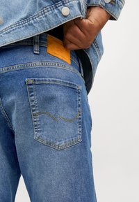 Lichtblauwe denim jeans met een oranje leren patch op de tailleband. Voorzien van een achterzak met gele stiksels en een gebogen ontwerpdetail.