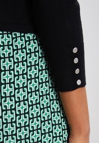 Bras portant une manche noire avec quatre boutons argentés, associé à un tissu à motif géométrique vert et blanc.