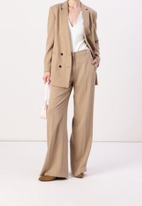 Beige maatpak met een dubbelgebreide blazer en wijde broekspijpen. Gekoşt in combinatie met een witte top en een delicate roze handtas. Bruinehakken.