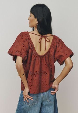 Woman with long dark hair wears a rust-red eyelet blouse with open back tied at the neck and blue denim jeans, hands on hips.