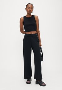 Schwarzes, kurzes Tanktop und weit geschnittene Hosen mit Knopfdetails an den Seiten. Das Model trägt Plateauschuhe und hält eine schwarze Handtasche.