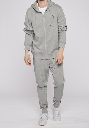 Homme portant un sweat à capuche zippé gris et un jogging assorti, se tenant devant un fond uni, avec des baskets blanches. Style de vêtements de sport décontracté.