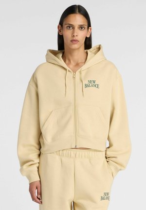 Vrouw draagt een beige New Balance hoodie met ritssluiting en bijpassende joggingbroek, staat met één hand in de zak tegen een effen achtergrond.
