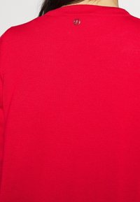 Roter Strickpullover mit glatter Textur und rundem Ausschnitt, ausgestattet mit einem kleinen silbernen Logo-Detail auf der Rückseite.