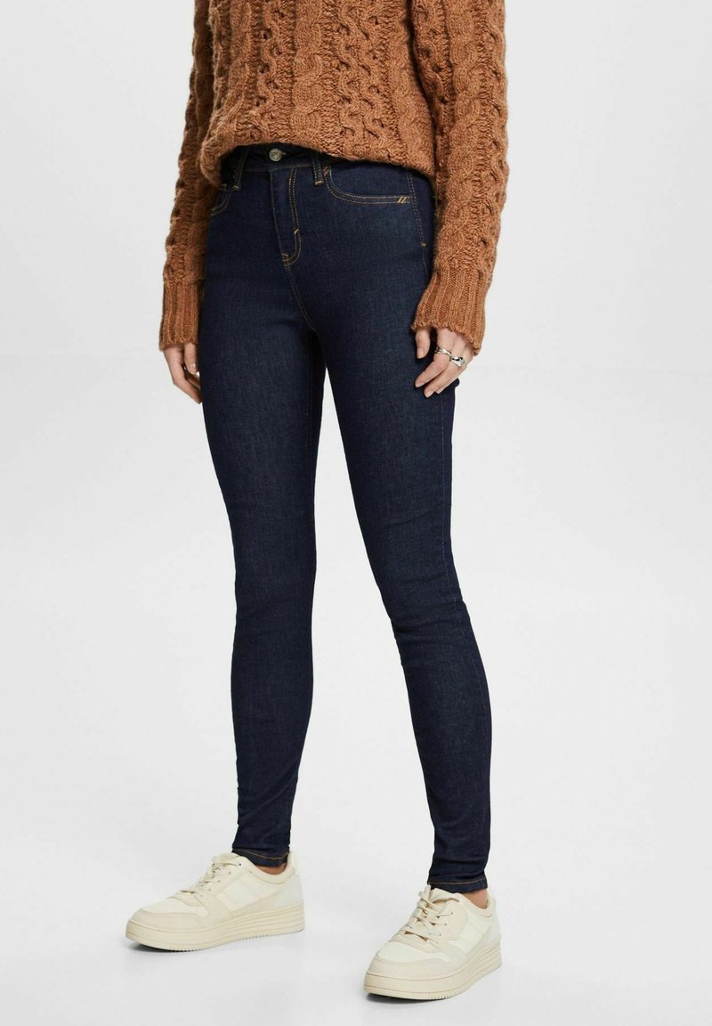 Jeans skinny de mezclilla azul oscuro con una cintura alta y una textura suave, combinados con un suéter de punto marrón y zapatillas blancas.