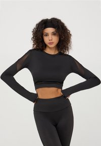 Top crop sportivo nero a maniche lunghe, con pannelli in rete. Tessuto liscio, design aderente che mette in risalto il girovita.