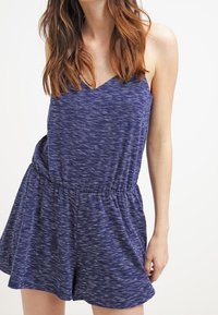 Rompers sans manches bleu avec un décolleté en V, une taille élastique et un tissu texturé, présentant un motif moucheté subtil. Design décontracté et léger.
