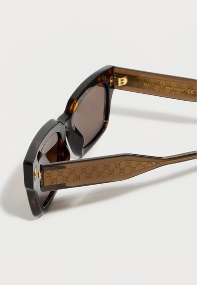 Γυαλιά ηλίου με τετράγωνο σκελετό σε σχέδιο χελώνας, με καφέ νατουραλιστικό μοτίβο Gucci στα μπράτσα και χρυσούς μεντεσέδες, απεικονίζονται από πλάγια γωνία σε λευκό φόντο.