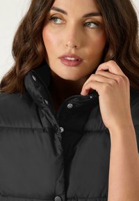 Gilet matelassé noir avec un col haut et des boutons-pression. Il possède une fermeture éclair et des détails en métal brillant, offrant une texture lisse.