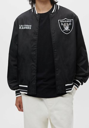 Osoba ubrana w czarną kurtkę varsity Las Vegas Raiders z paskowanymi mankietami i kołnierzem oraz białe spodnie, stojąca na tle jednolitego tła.