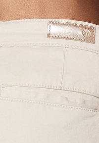 Tejido de denim beige con una textura suave, que presenta una etiqueta metálica con un logo en relieve y un único bolsillo trasero. La costura es visible.