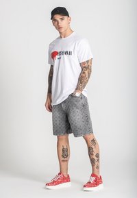 Bijela majica s crnim natpisom i crvenim akcentskim detaljima, sivi kratki hlače sa uzorkom, crvene tenisice s točkicama i vidljive tetovaže na rukama i nogama.